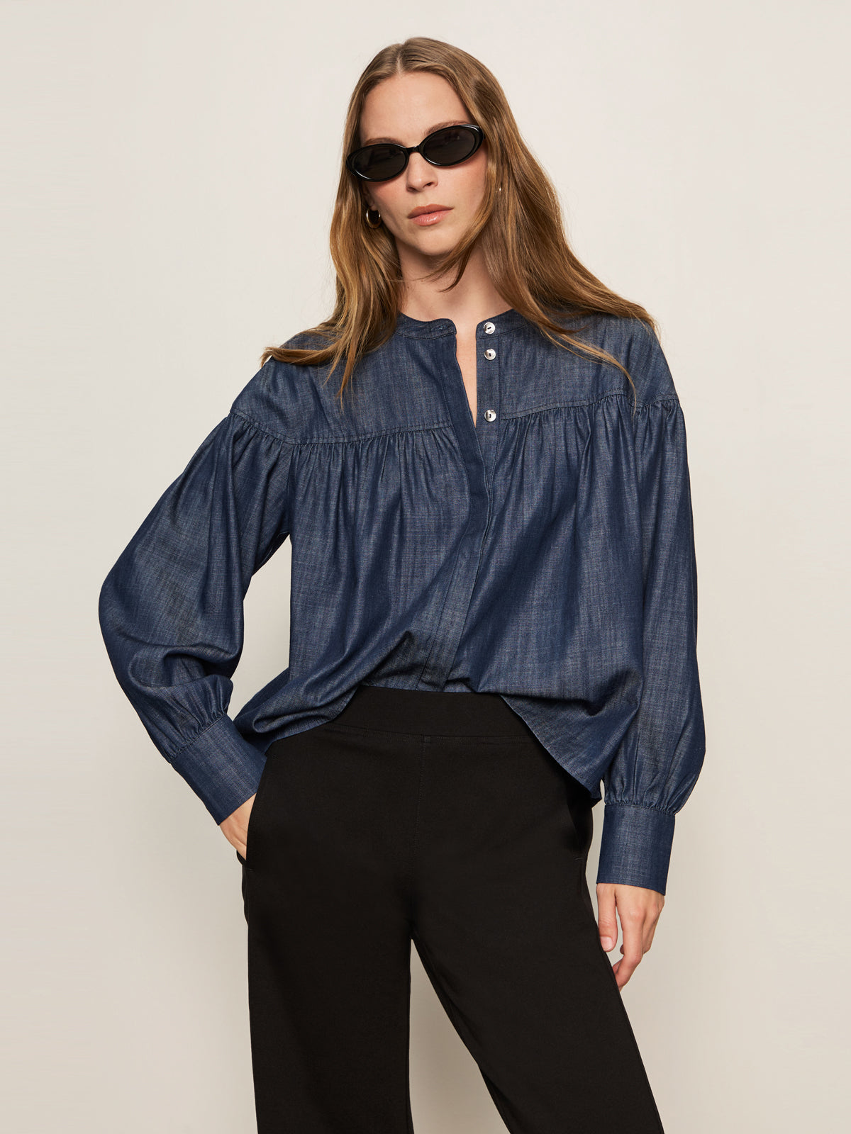 A woman with long brown hair wears black sunglasses, black pants, and the Sanctuary Clothing personal best blouse moonshadow—a loose-fitting blue denim button-up—standing against a plain light background with one hand in her pocket.