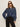 A woman with long brown hair wears black sunglasses, black pants, and the Sanctuary Clothing personal best blouse moonshadow—a loose-fitting blue denim button-up—standing against a plain light background with one hand in her pocket.