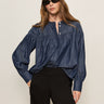 A woman with long brown hair wears black sunglasses, black pants, and the Sanctuary Clothing personal best blouse moonshadow—a loose-fitting blue denim button-up—standing against a plain light background with one hand in her pocket.