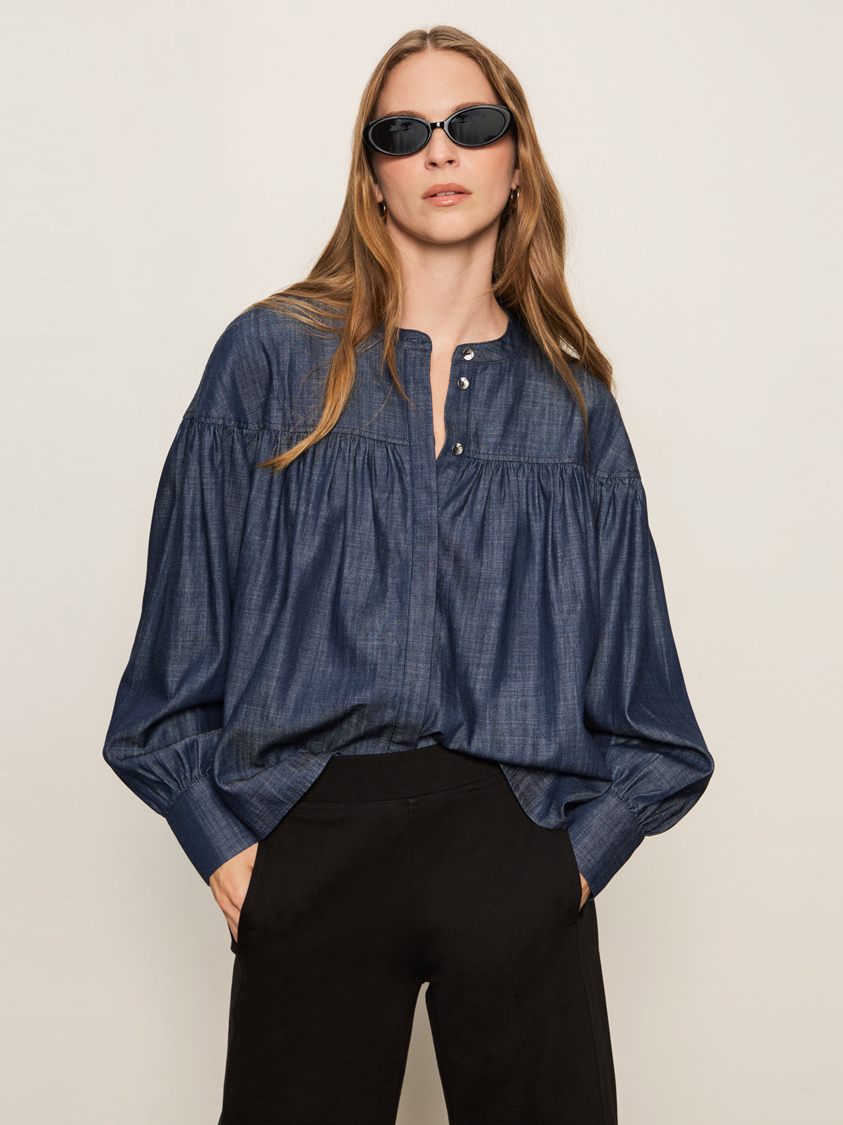 A woman with long light brown hair wears black sunglasses, black pants, and the Sanctuary Clothing personal best blouse in moonshadow. She stands against a plain light background with her hands in her pockets.