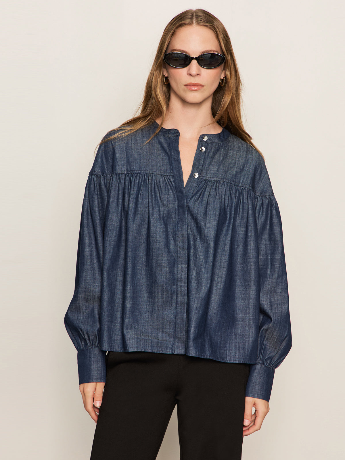 A woman in Sanctuary Clothing’s personal best blouse moonshadow—a loose, long-sleeve denim top with a gathered yoke and buttoned collar—pairs it with black pants, standing against a plain light background.