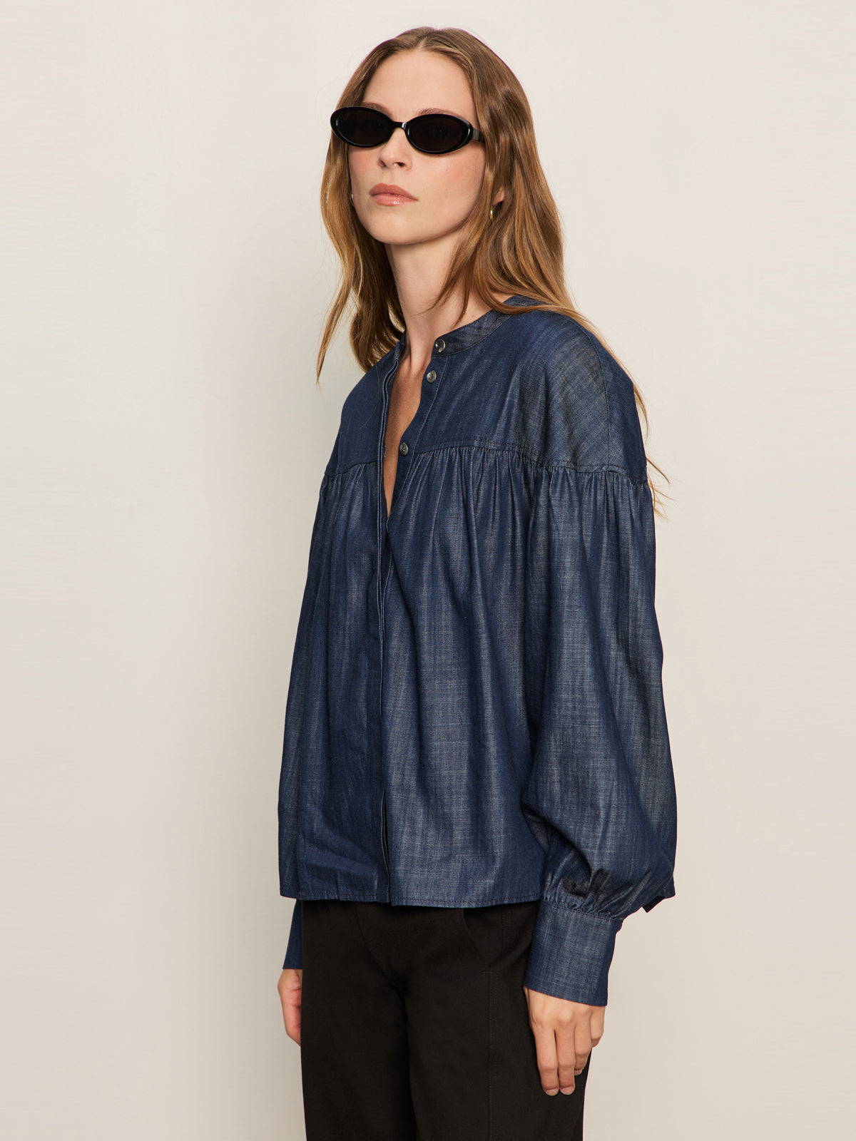 A woman with long, light brown hair models the Sanctuary Clothing personal best blouse moonshadow—black sunglasses, black pants, and a loose-fitting, dark denim top—standing against a plain light background.
