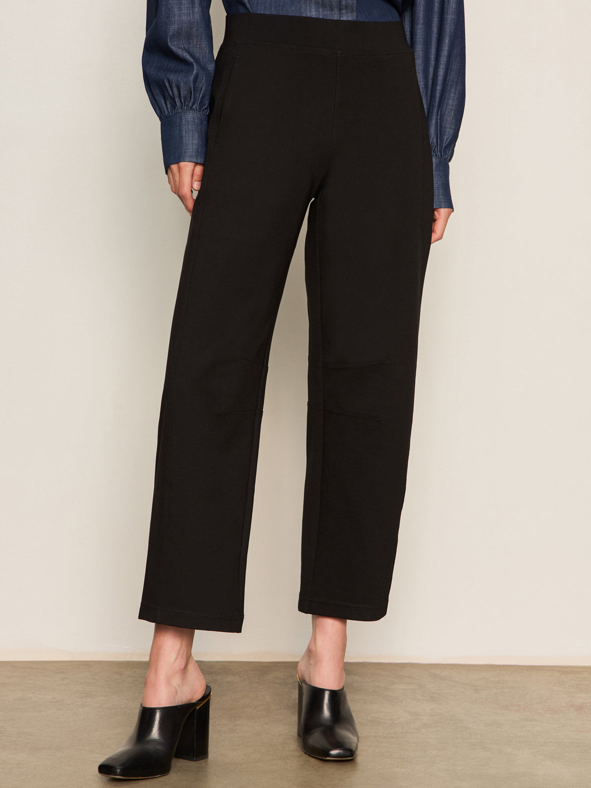 A person models the Sanctuary Clothing ponte barrel trouser in black, paired with a blue long-sleeve shirt and black heeled mules, standing against a plain light background. The waist-up photo excludes the person's face.
