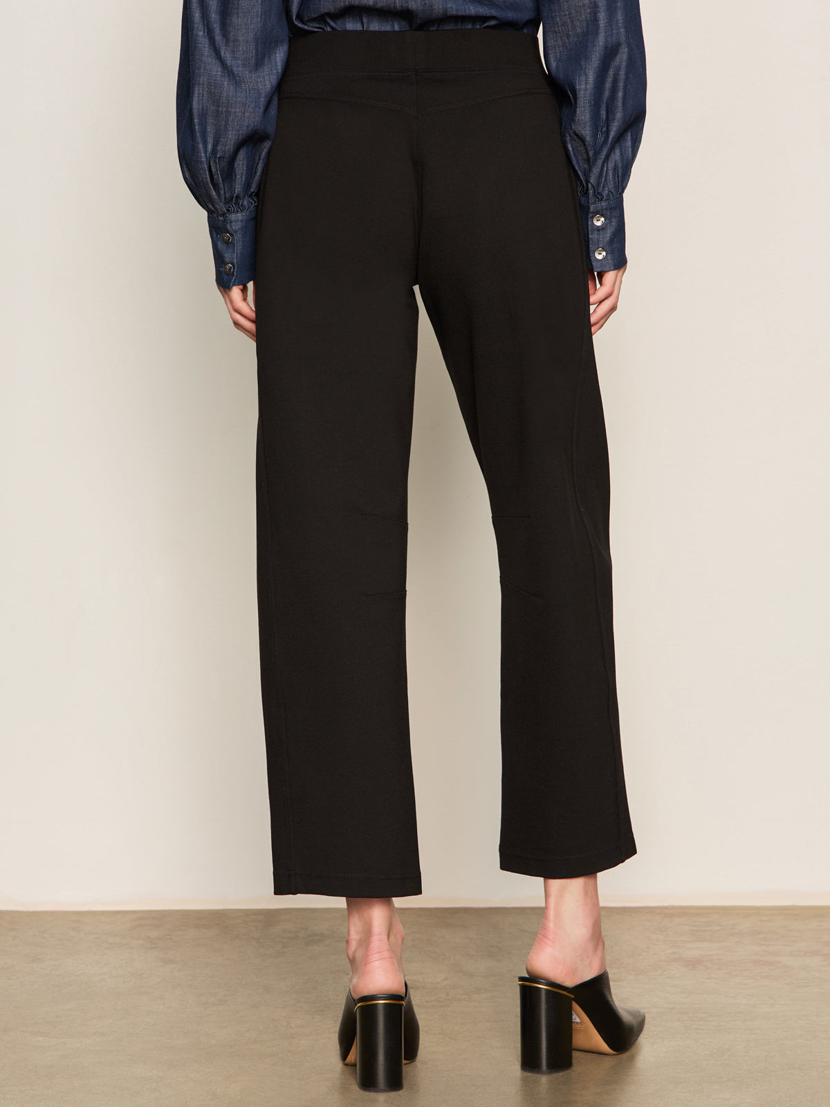 A person wearing Sanctuary Clothing's ponte barrel trouser in black, shown from the back indoors against a neutral background, styled with a dark blue long-sleeve blouse and black heeled mules.