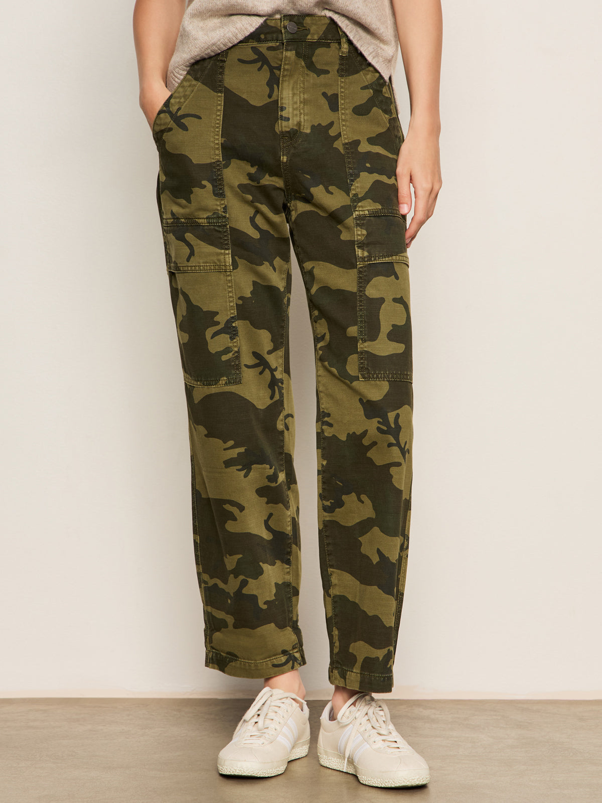A person models the Sanctuary Clothing boyfriend utility pant in rambler camo, featuring side pockets and a relaxed fit, paired with white sneakers and a beige top. Only the lower half is visible against a plain background.