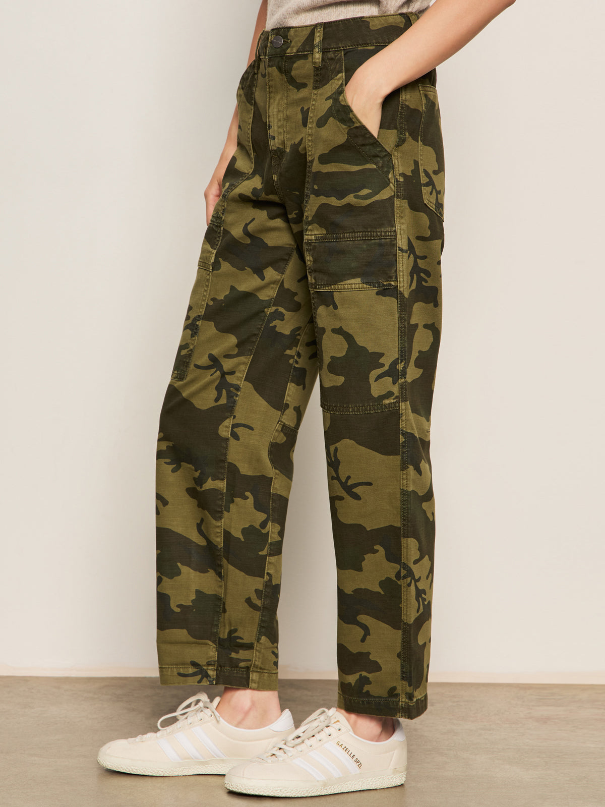 Person wearing Sanctuary Clothing boyfriend utility pant rambler camo, paired with white sneakers. Only the lower half of the body is visible, standing on a light floor against a plain beige wall.