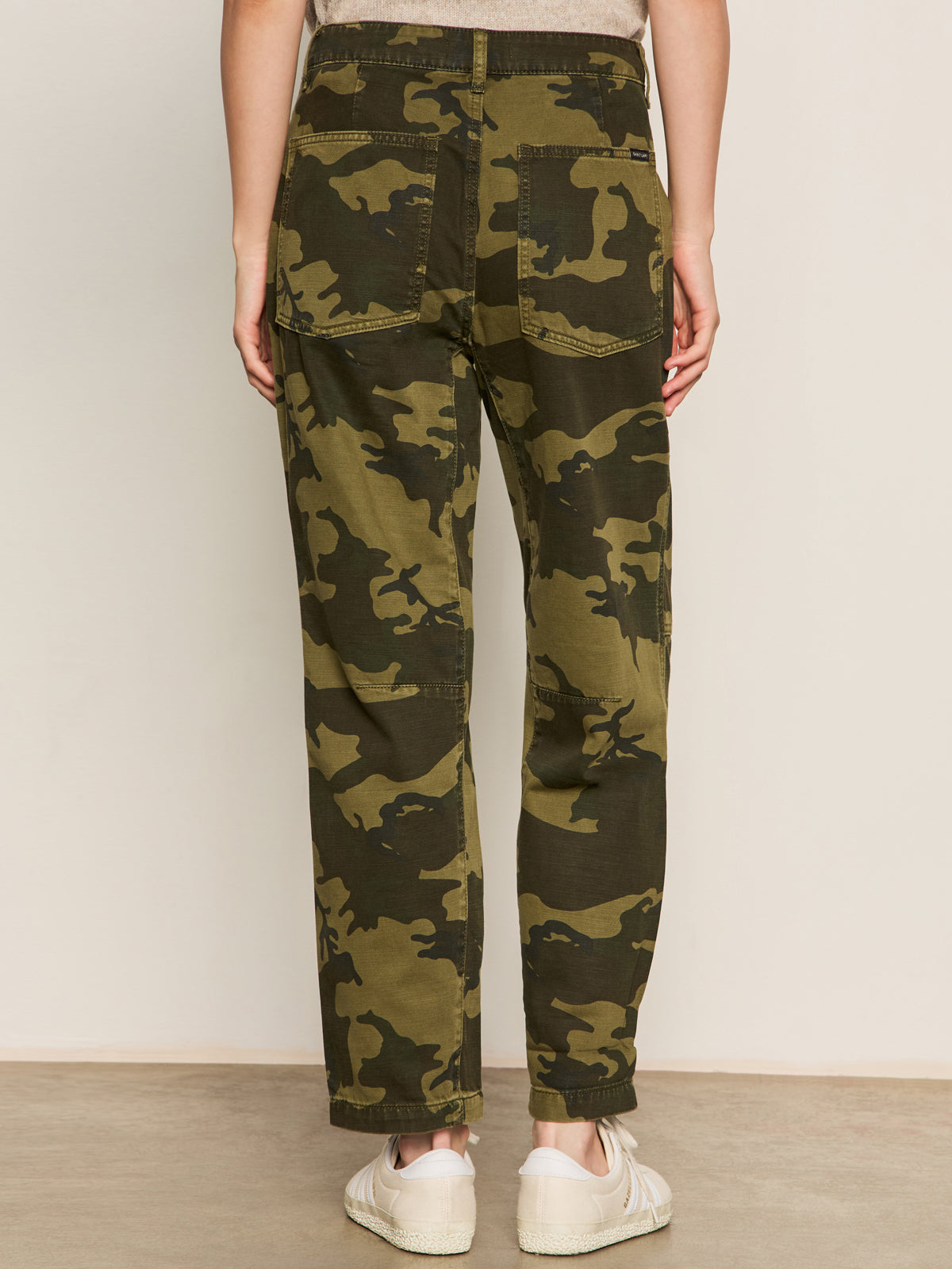 A model faces away to show the back pockets of the Sanctuary Clothing boyfriend utility pant in rambler camo, paired with white sneakers, against a plain background.