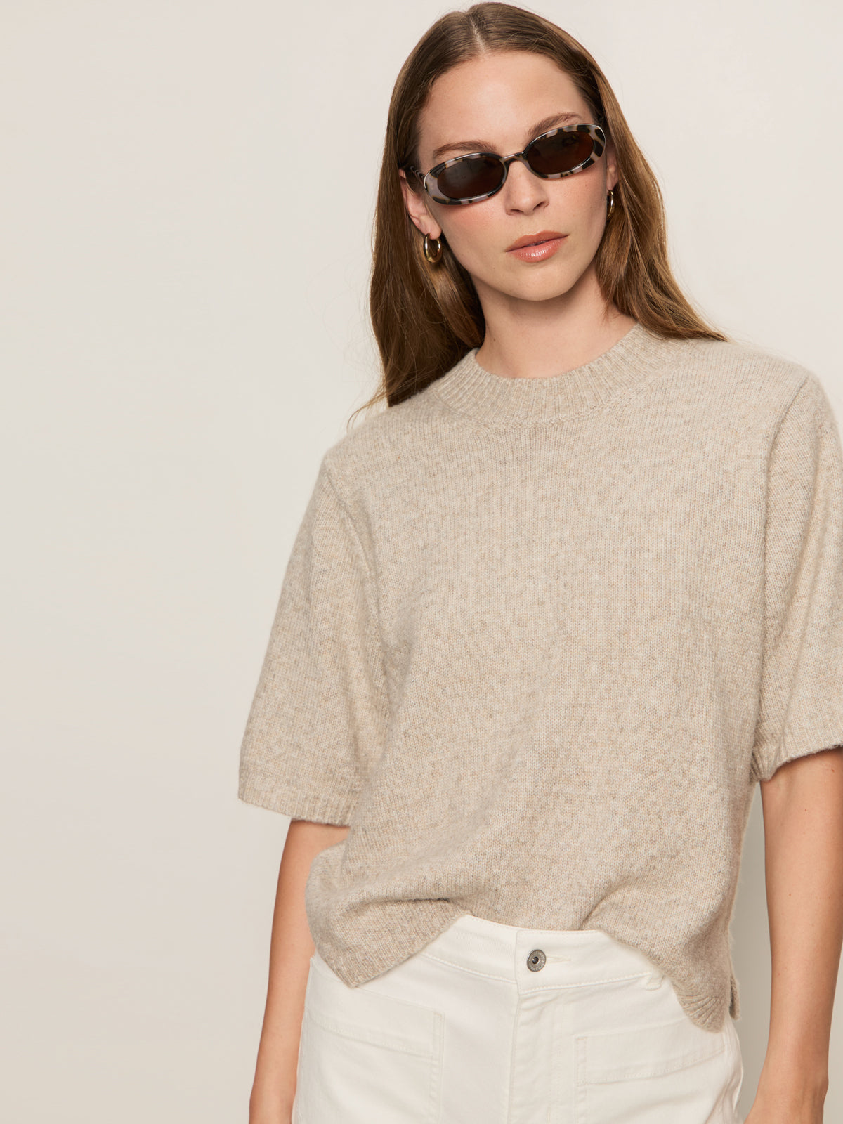 A woman with long brown hair wears black sunglasses, white pants, and the Sanctuary Clothing perfect sweater tee in heather pearl. She stands against a plain light background, looking slightly to the side with a neutral expression.