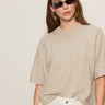 A woman with long brown hair wears black sunglasses, white pants, and the Sanctuary Clothing perfect sweater tee in heather pearl. She stands against a plain light background, looking slightly to the side with a neutral expression.