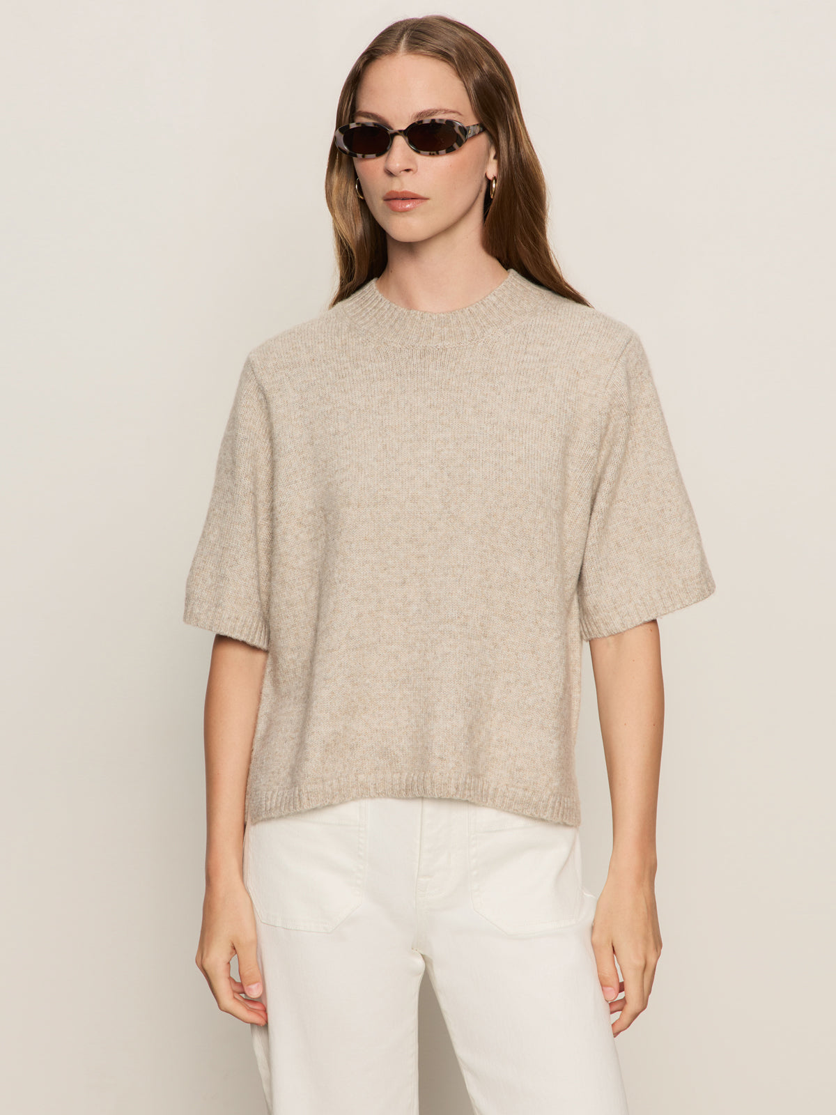 A woman with long brown hair wears tortoiseshell sunglasses, white pants, and the Sanctuary Clothing perfect sweater tee in heather pearl, standing against a plain light background.