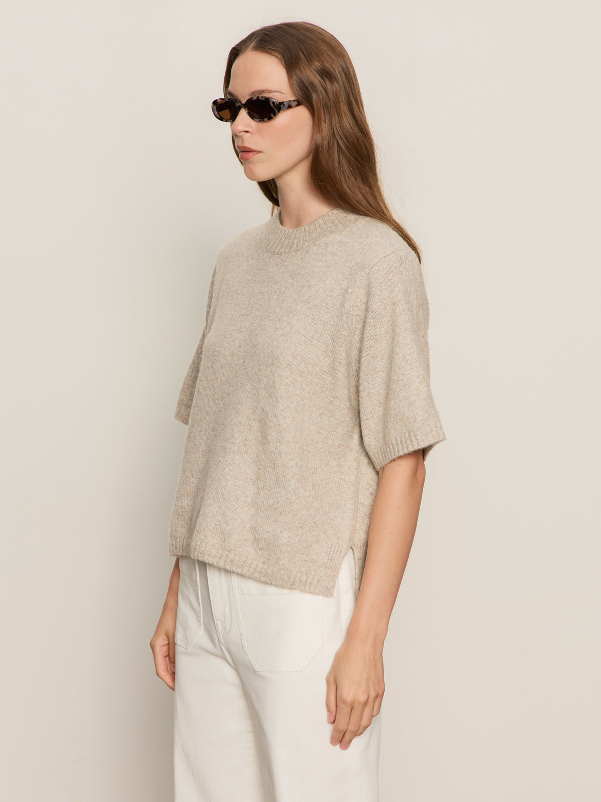 A woman with long brown hair wears dark sunglasses, white pants, and the Sanctuary Clothing perfect sweater tee in heather pearl, standing against a plain light background.