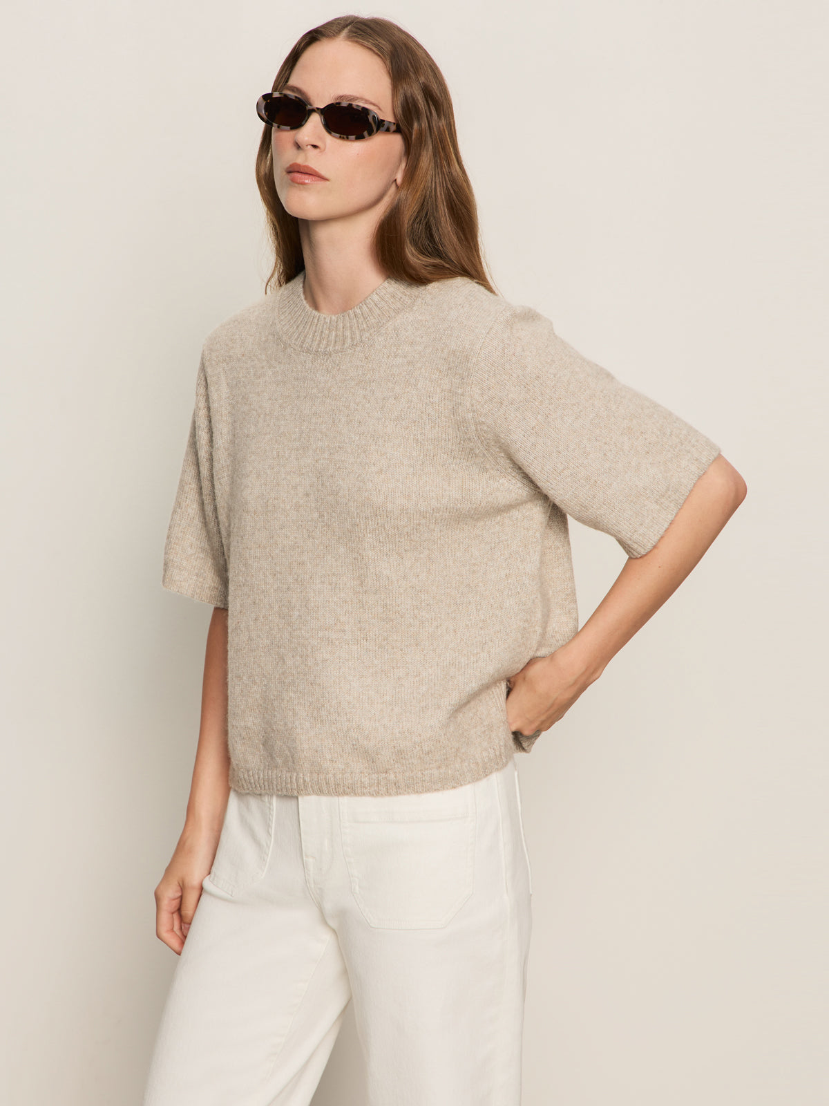 A woman with long brown hair, wearing dark sunglasses and the Sanctuary Clothing perfect sweater tee in heather pearl, pairs it with white pants as she poses against a plain light background, one hand on her hip.
