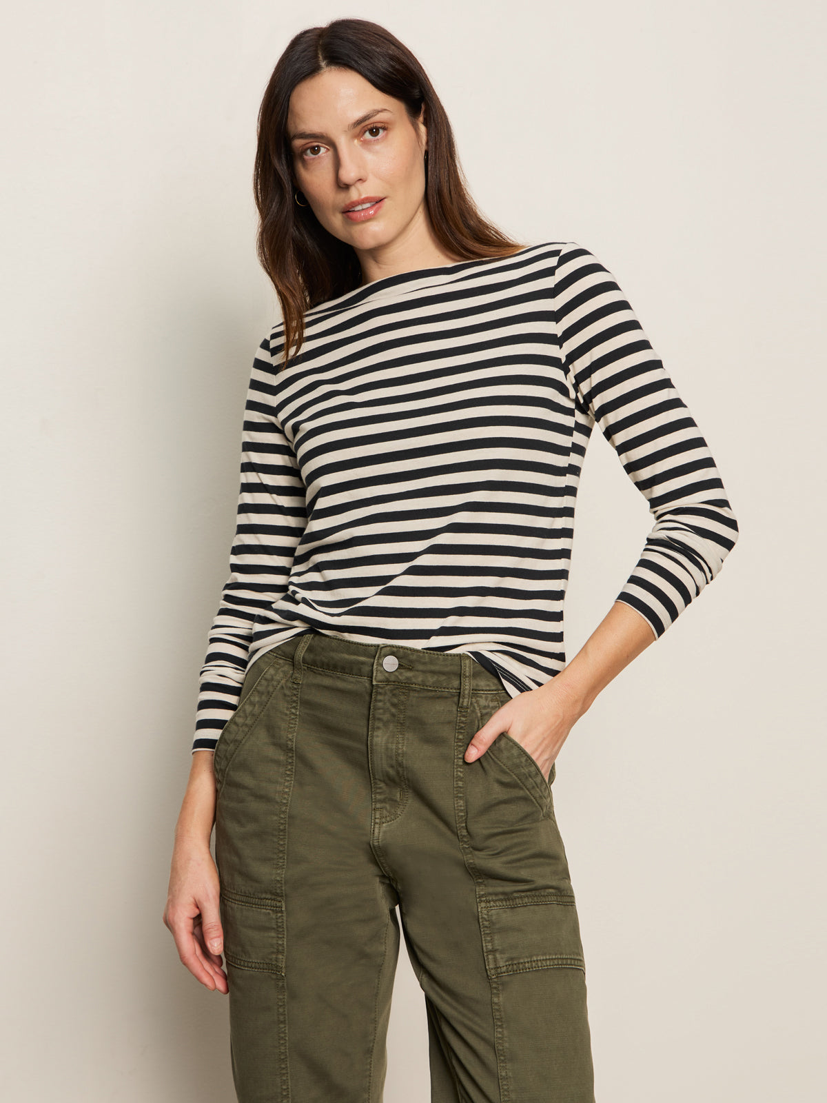 A woman poses against a light background wearing the Sanctuary Clothing boatneck long sleeve tee in light oat black stripe, paired with olive green cargo pants. She has a neutral expression and one hand in her pocket.