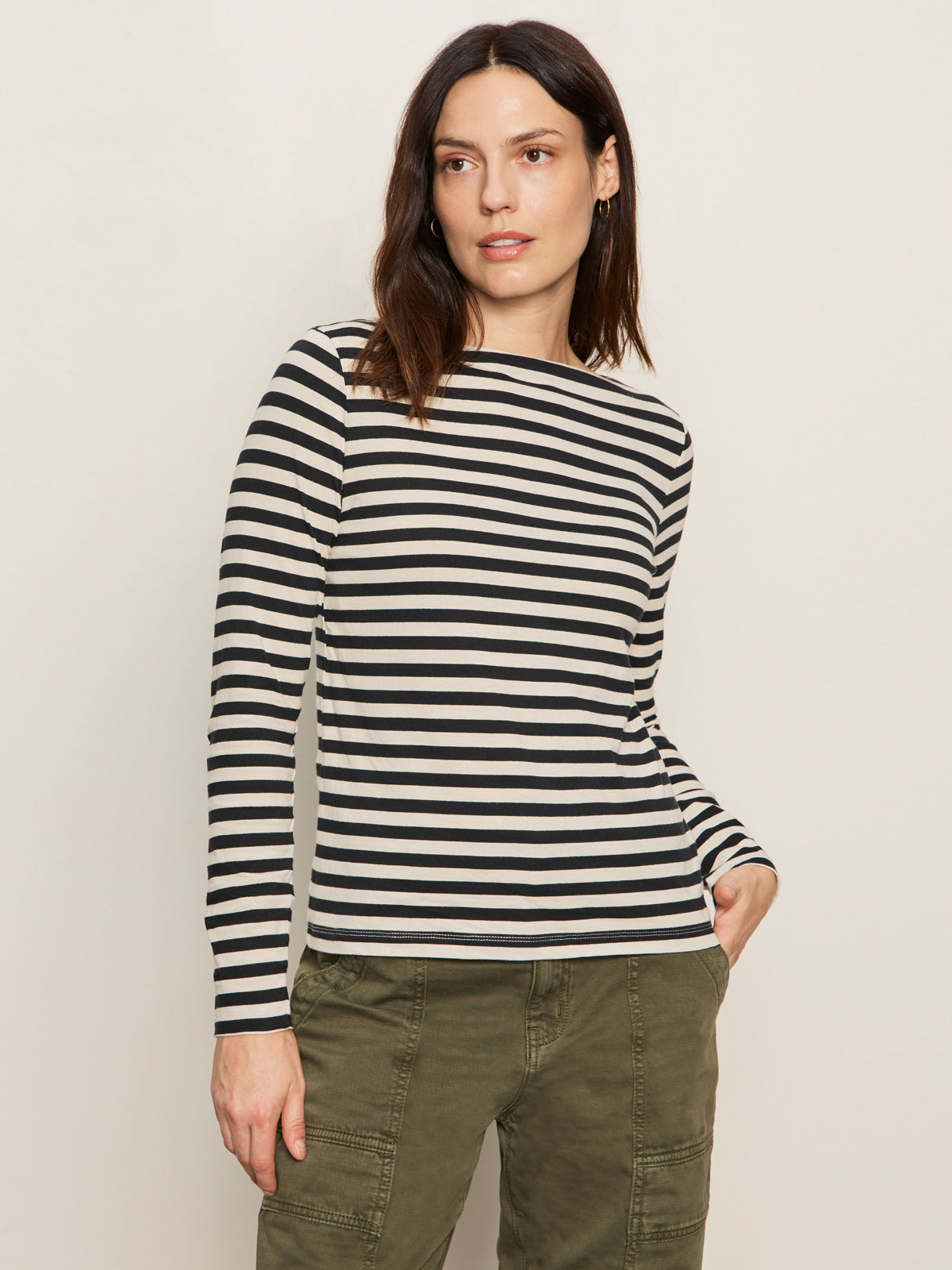 A woman models the Sanctuary Clothing boatneck long sleeve tee in light oat black stripe, standing against a plain background and looking slightly to the side with one hand in her pocket.