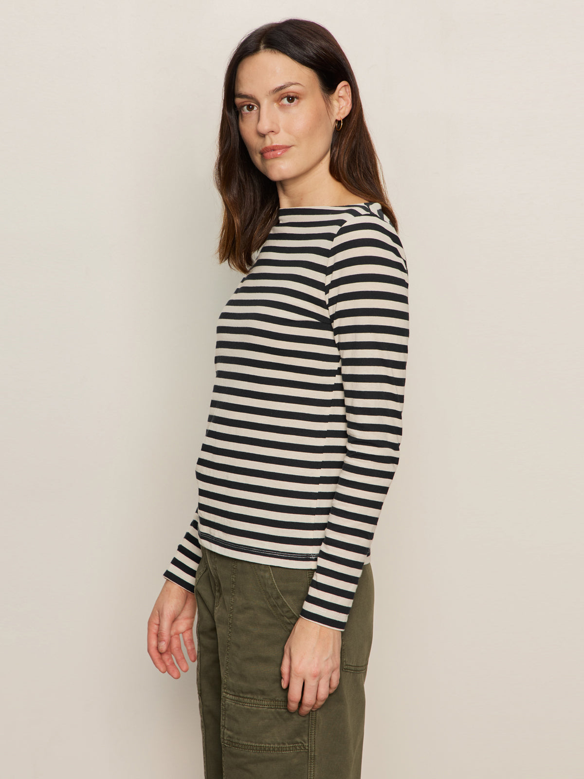 A woman with long brown hair wears the Sanctuary Clothing boatneck long sleeve tee in light oat black stripe 3 and stands sideways against a plain light background.