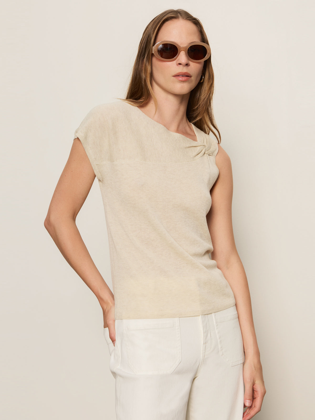 A woman with long straight hair wears the Sanctuary Clothing asymm shoulder twist top in heather pearl and white pants, paired with round brown sunglasses. She poses with one hand in her pocket against a plain light background.