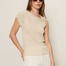 A woman with long straight hair wears the Sanctuary Clothing asymm shoulder twist top in heather pearl and white pants, paired with round brown sunglasses. She poses with one hand in her pocket against a plain light background.