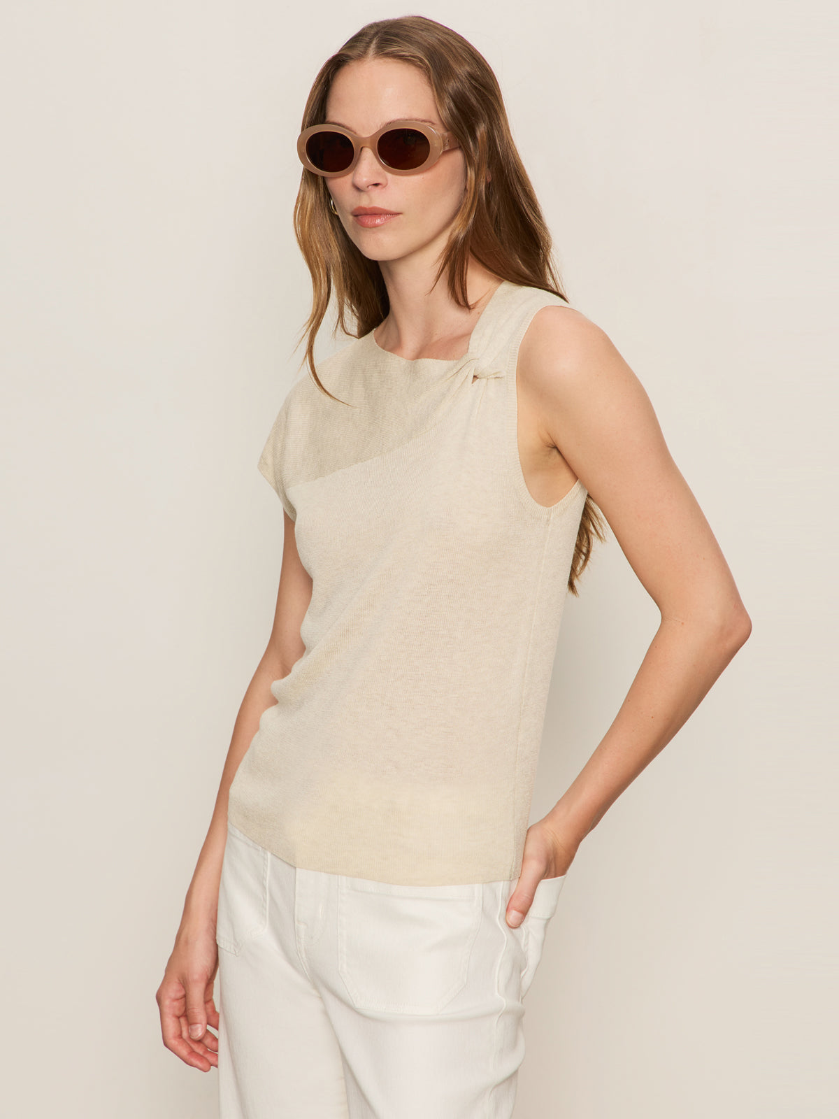 A woman with long brown hair wears round sunglasses and the Sanctuary Clothing asymm shoulder twist top in heather pearl, paired with white pants, standing against a plain light background.
