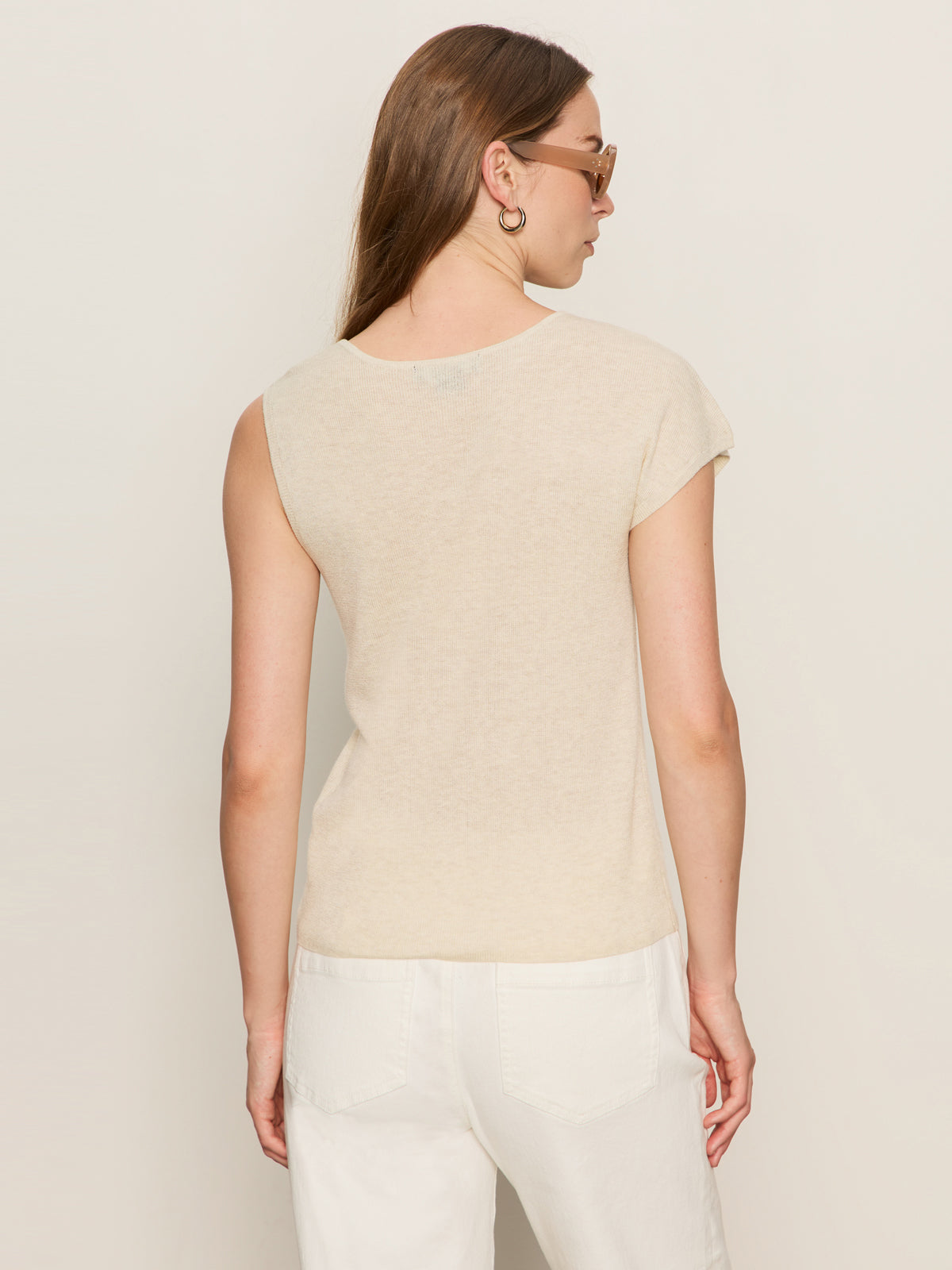 A woman with long brown hair, sunglasses, and hoop earrings is seen from behind wearing the Sanctuary Clothing asymm shoulder twist top in heather pearl with white pants against a plain light background.