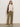 A woman stands against a plain wall wearing the Sanctuary Clothing draped sleeveless mock neck top in mother of pearl, olive wide-leg pants, white sneakers, and a brown shoulder bag. She has long brown hair and her hands in her pockets.