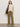 A woman with long hair and a neutral expression stands against a plain wall wearing the Sanctuary Clothing draped sleeveless mock neck top in mother of pearl, olive green wide-leg pants, cream sneakers, and a dark brown shoulder bag.