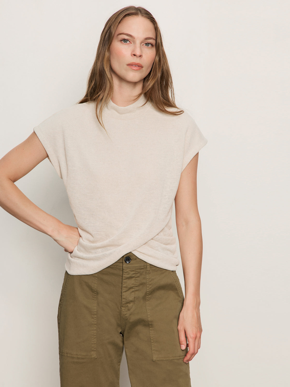A woman with long brown hair wears the Sanctuary Clothing draped sleeveless mock neck top in mother of pearl, paired with olive green high-waisted pants, standing against a plain light-colored wall with one hand on her hip.