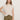 A woman with long brown hair wears the Sanctuary Clothing draped sleeveless mock neck top in mother of pearl, paired with olive green high-waisted pants, standing against a plain light-colored wall with one hand on her hip.