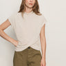 A woman with long brown hair wears the Sanctuary Clothing draped sleeveless mock neck top in mother of pearl, paired with olive green high-waisted pants, standing against a plain light-colored wall with one hand on her hip.