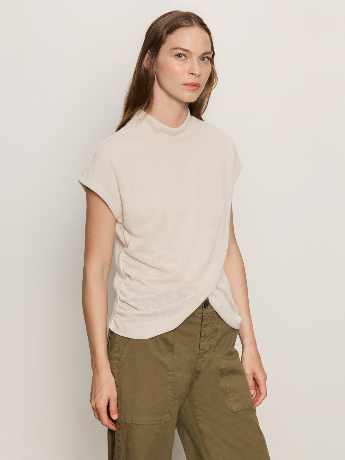 A woman with long brown hair wears the Sanctuary Clothing draped sleeveless mock neck top in mother of pearl, paired with olive green pants. She stands against a plain light background, looking slightly to the side with a neutral expression.