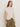 A woman with long brown hair wears the Sanctuary Clothing draped sleeveless mock neck top in mother of pearl, paired with olive green pants. She stands against a plain light background, looking slightly to the side with a neutral expression.