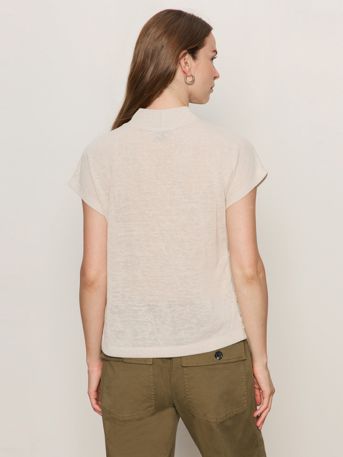 A woman with long brown hair stands with her back to the camera, wearing the Sanctuary Clothing draped sleeveless mock neck top in mother of pearl, paired with olive green pants against a plain light background.