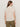 A woman with long brown hair stands with her back to the camera, wearing the Sanctuary Clothing draped sleeveless mock neck top in mother of pearl, paired with olive green pants against a plain light background.