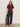 A woman stands against a plain wall wearing the Sanctuary Clothing shirred waist dolman tee in chicory, paired with dark blue wide-leg jeans and white sneakers. She holds a leopard print jacket and has straight, light brown hair.
