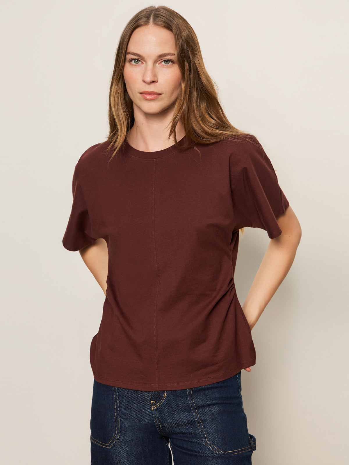 A woman with long, light brown hair wears the Sanctuary Clothing shirred waist dolman tee in chicory and dark blue jeans, standing against a plain light background with her hands in her pockets.