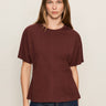 A woman with straight, light brown hair wears the Sanctuary Clothing Shirred Waist Dolman Tee in chicory and dark jeans, standing against a beige background and looking at the camera with a neutral expression.