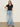 A woman with long hair poses against a plain light wall, wearing Sanctuary Clothing's notch neck tee in black, light blue wide-leg jeans with a black belt, and black sandals. One hand rests on her hip.