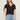 A woman with long, light brown hair wears Sanctuary Clothing’s notch neck tee in black, tucked into light blue jeans with a black belt. She stands against a plain, light-colored background, looking slightly past the camera.