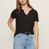 A woman with long, light brown hair wears Sanctuary Clothing’s notch neck tee in black, tucked into light blue jeans with a black belt. She stands against a plain, light-colored background, looking slightly past the camera.