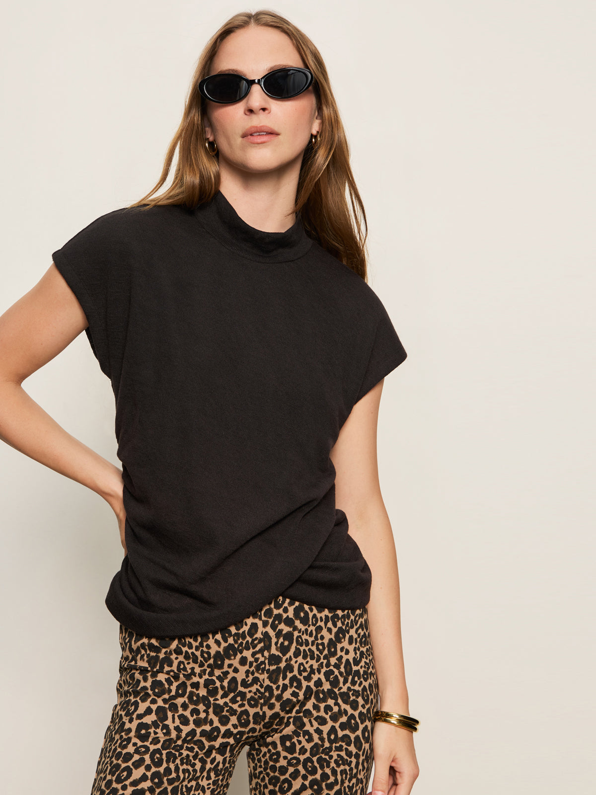 A woman poses confidently with one hand on her hip against a plain light background, wearing black sunglasses, leopard print pants, and the Sanctuary Clothing draped sleeveless mock neck top in black.