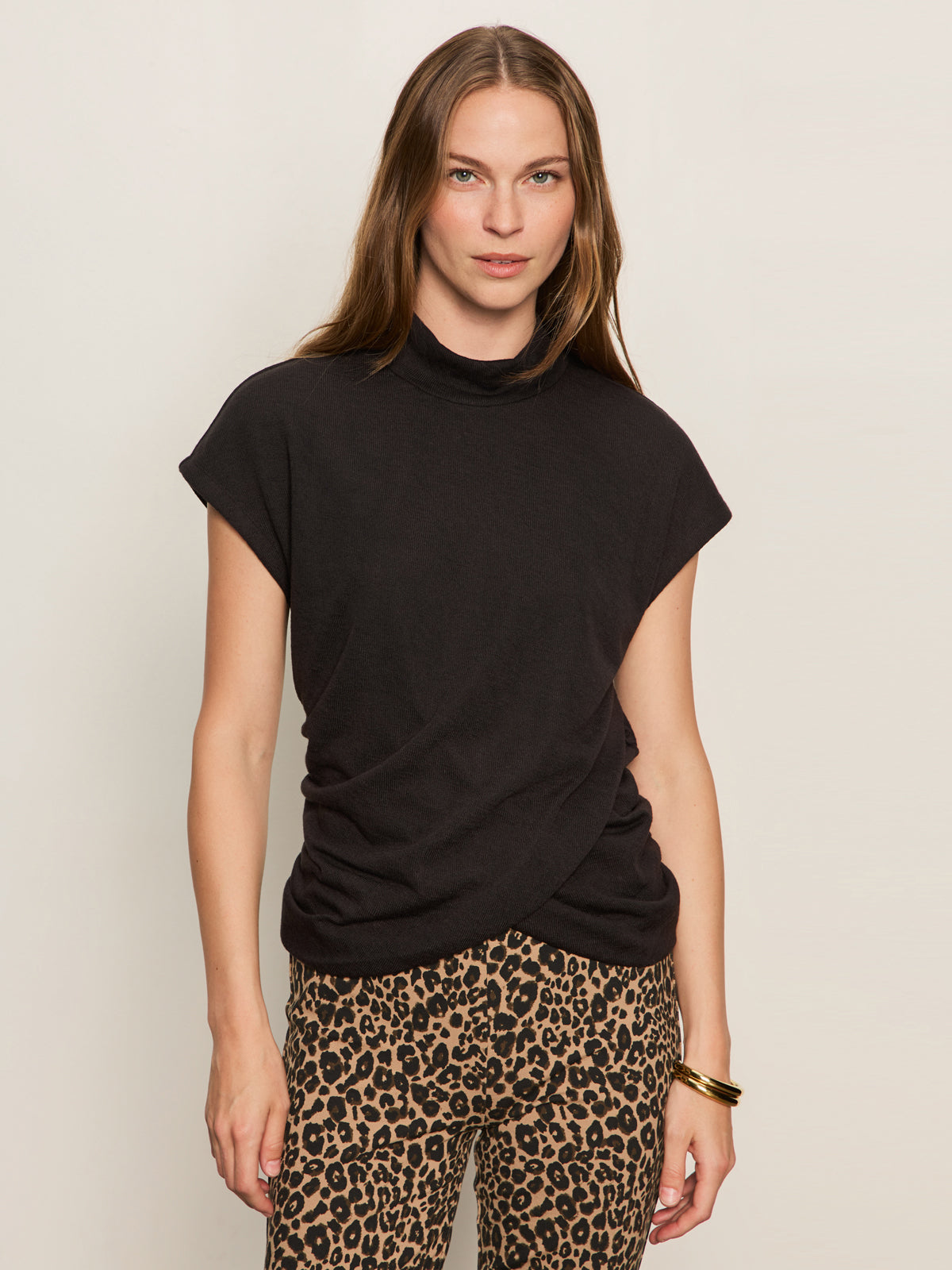A woman with long brown hair models the Sanctuary Clothing draped sleeveless mock neck top in black, paired with leopard-print pants and gold bangles, standing against a plain light background with a neutral expression.