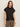 A woman with long brown hair models the Sanctuary Clothing draped sleeveless mock neck top in black, paired with leopard-print pants and gold bangles, standing against a plain light background with a neutral expression.