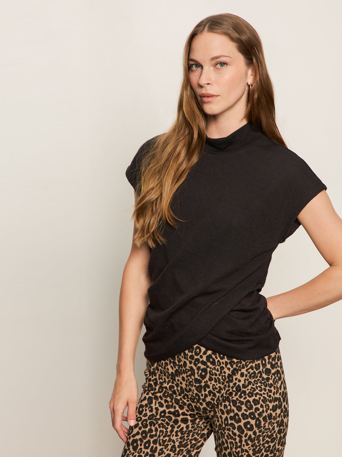 A woman with long, light brown hair wears the Sanctuary Clothing draped sleeveless mock neck top in black and leopard print pants, standing against a plain light background with one hand on her hip.