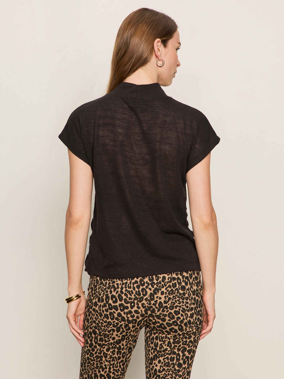 A woman with long brown hair, facing away, wears the Sanctuary Clothing draped sleeveless mock neck top in black and high-waisted leopard print pants, accessorized with gold earrings and a bracelet, against a plain light background.