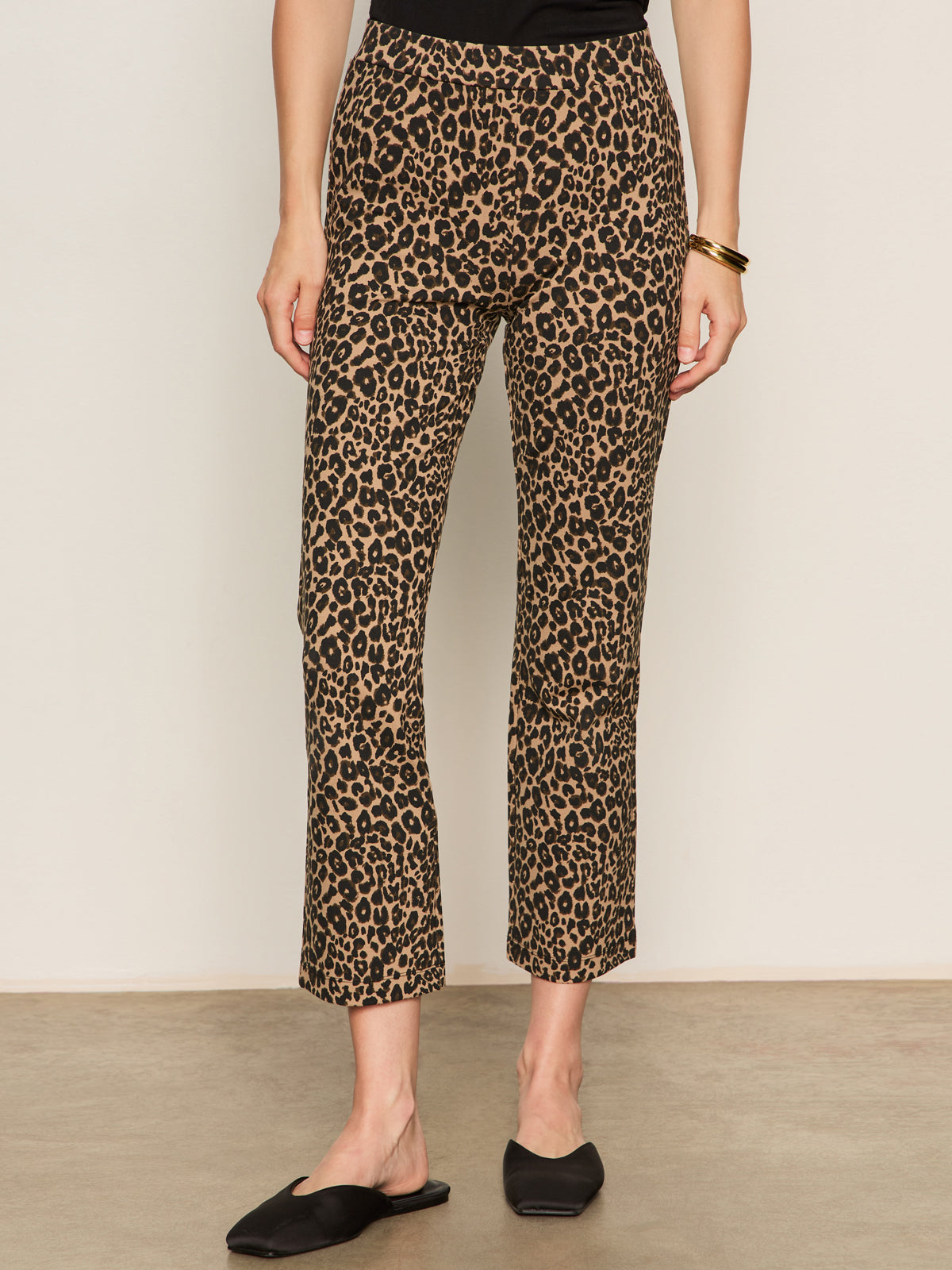 A person wears Sanctuary Clothing's Carnaby Kick Crop Leggings in French Kitty print, paired with a black top, black slip-on shoes, and a gold bangle, standing relaxed against a beige wall.
