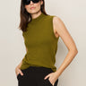 A woman with straight brown hair wears the "fuzzy shell martini olive" sleeveless turtleneck sweater by Sanctuary Clothing, paired with black sunglasses and pants, standing against a plain light background with hands in her pockets.