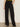 A person indoors wears Sanctuary Clothing's modern gab trouser black with a green top and black pointed-toe shoes, one hand in their pocket. Their head is not visible in the image.
