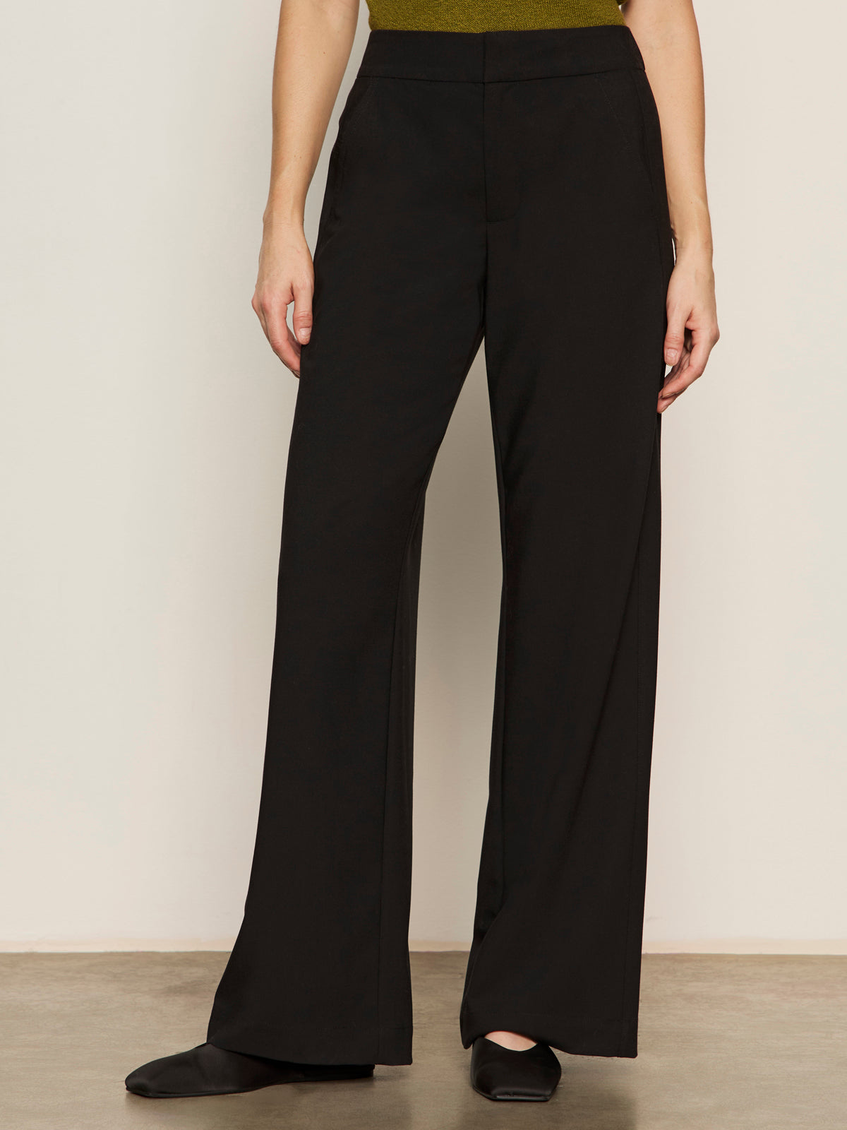 A person wearing Sanctuary Clothing’s modern gab trouser black with black pointed-toe shoes stands against a plain light-colored wall; only the lower half of the body is visible.
