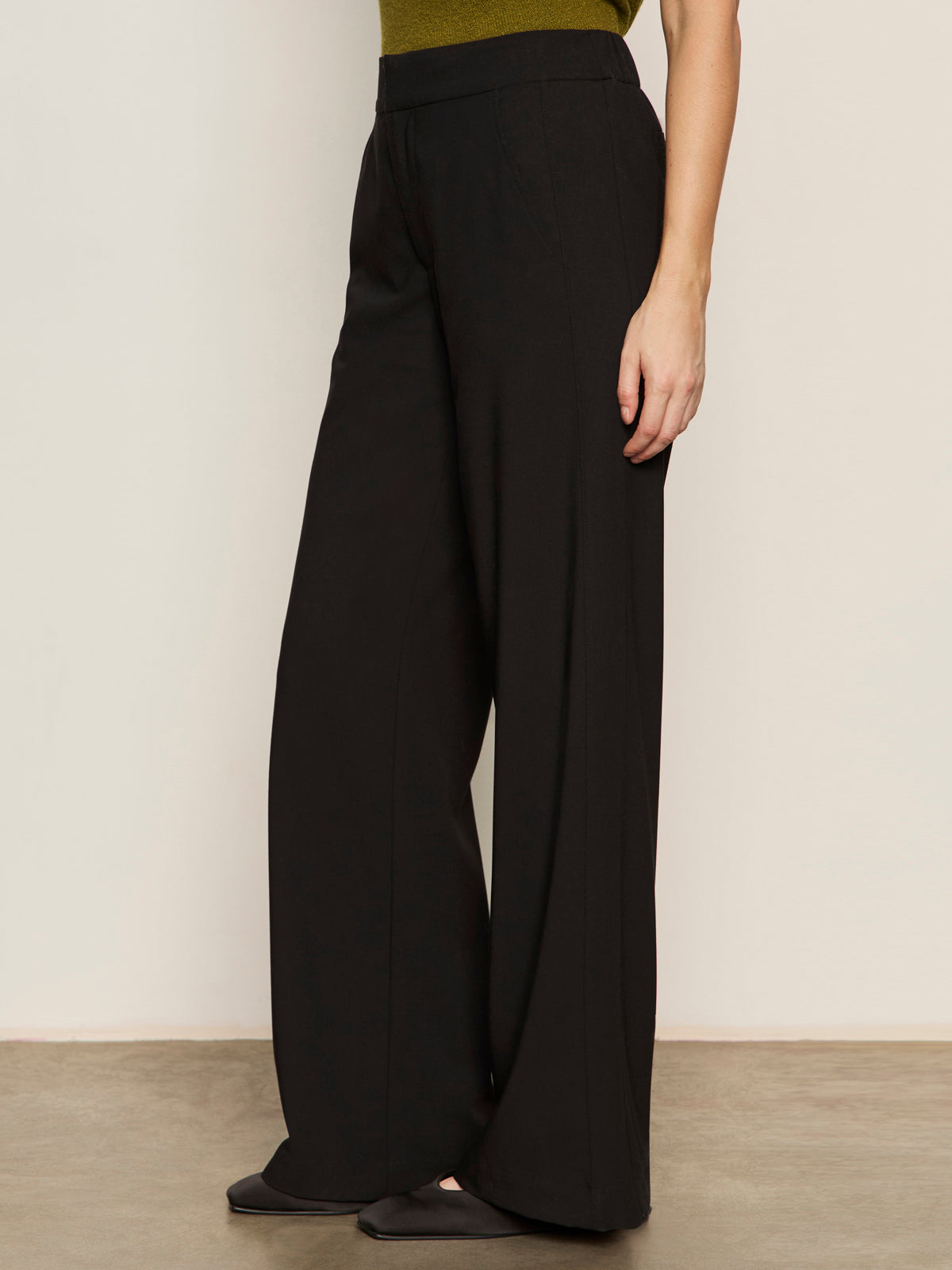 A person wearing Sanctuary Clothing's modern gab trouser in black and black flats stands against a light wall, with only the lower half of their body visible and their arm by their side.