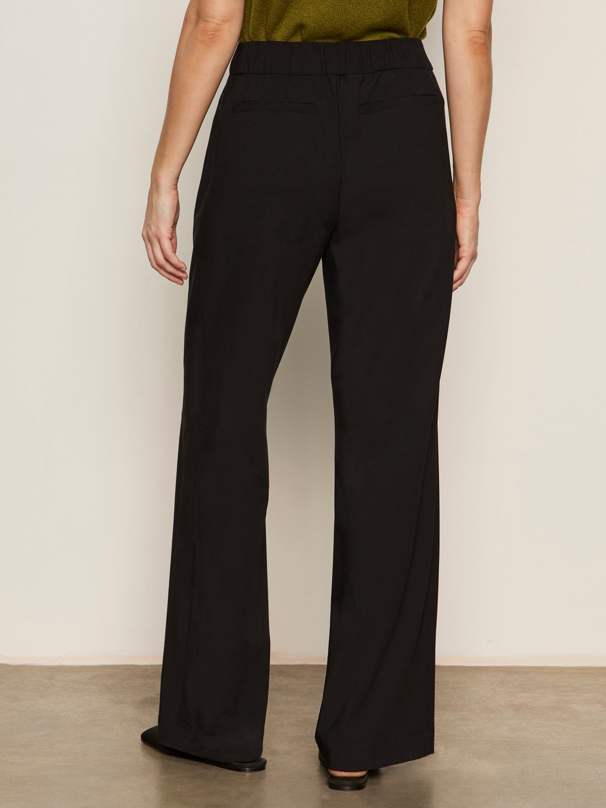 A person stands on a smooth floor facing away from the camera, wearing Sanctuary Clothing’s modern gab trouser black—high-waisted, wide-leg pants with a relaxed fit and elastic waistband—paired with a green top.