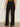 A person stands on a smooth floor facing away from the camera, wearing Sanctuary Clothing’s modern gab trouser black—high-waisted, wide-leg pants with a relaxed fit and elastic waistband—paired with a green top.